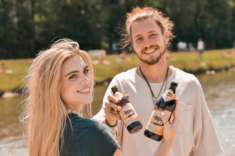 Zwei junge Erwachsene stehen lächelnd am Ufer eines Gewässers an einem sonnigen Tag. Beide halten jeweils eine Flasche Bazi-Cola-Mix in der Hand und stoßen an. Im Hintergrund sieht man eine grüne Wiese mit weiteren Personen.