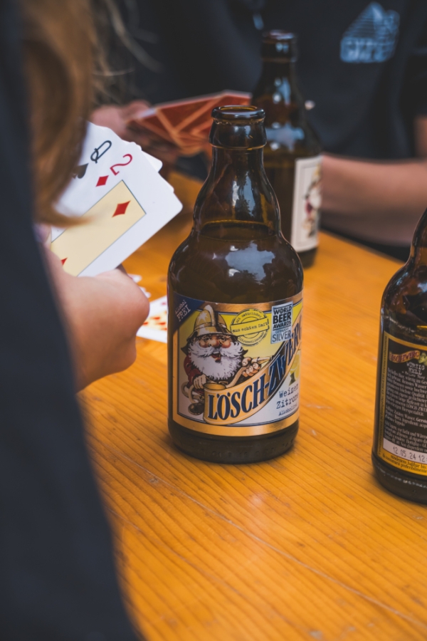 Eine Person spielt Karten an einem Holztisch, auf dem mehrere Flaschen Lösch-Zwerg stehen. Der Fokus liegt auf der Flasche Lösch-Zwerg Weizen Zitrone.
