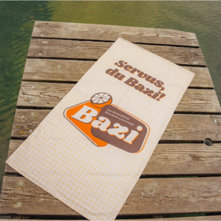 Beiges Bazi-Handtuch mit dem Schriftzug "Servus, du Bazi!" und großem Bazi-Logo liegt ausgebreitet auf einem Holzsteg am Wasser. Der Steg befindet sich über grünlich schimmerndem See.